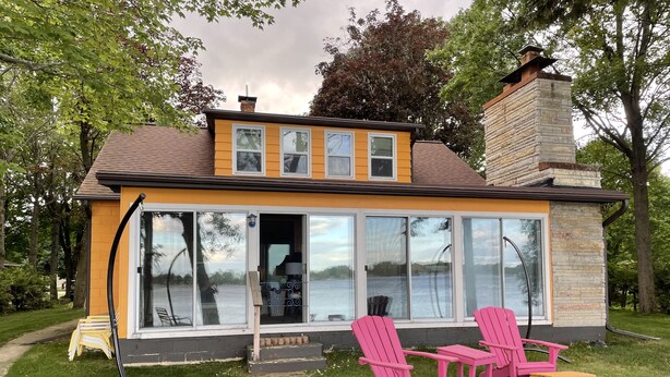 Une maison avec une grande porte vitrée, une cheminée en pierre et une cour verdoyante avec des chaises roses et une petite table.