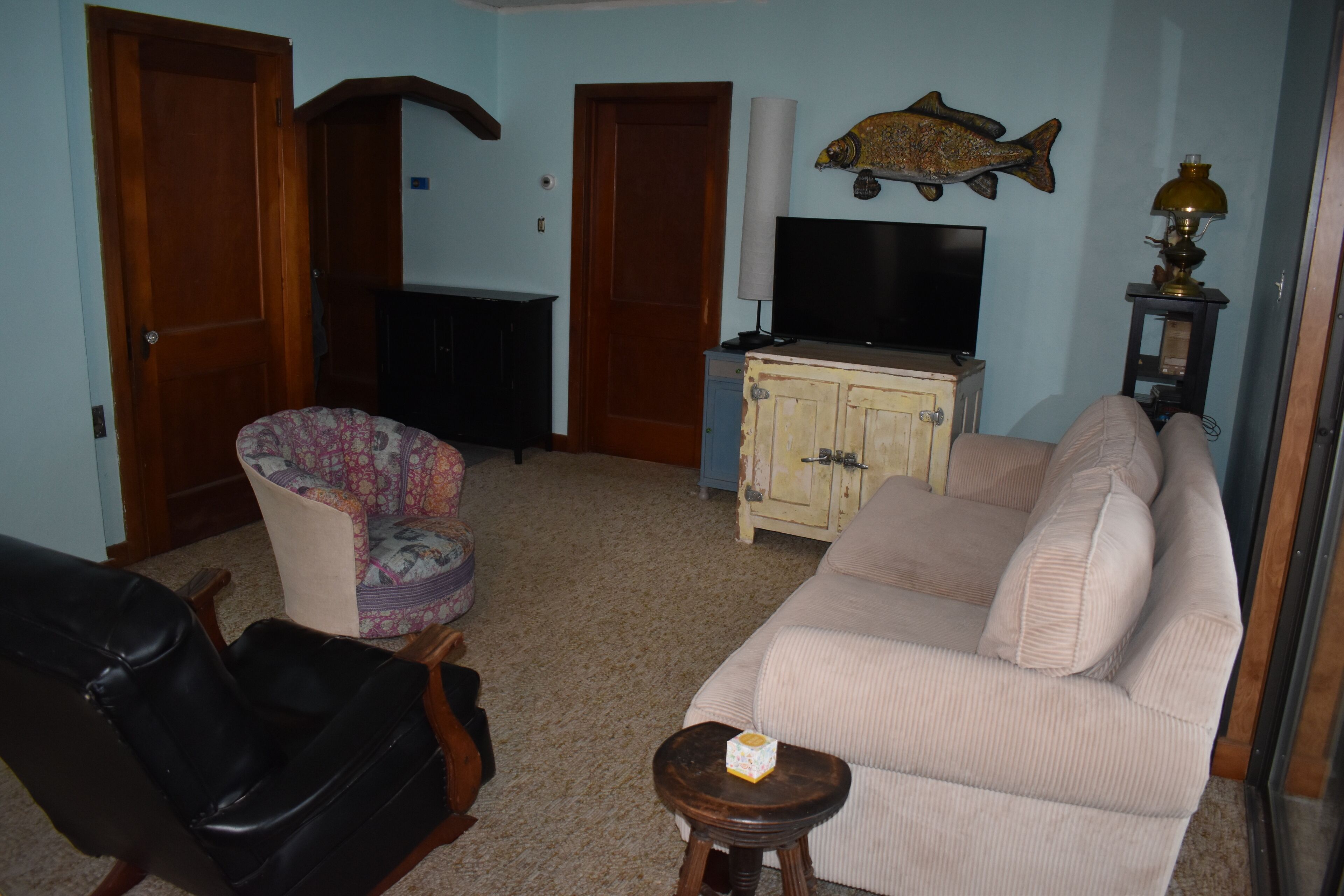 Una sala de estar con un sofá color beige, un sillón estampado, una butaca de cuero negro, un taburete de madera y un televisor sobre un mueble. Se observa una decoración con forma de pez en la pared.