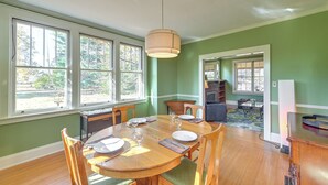 Un comedor con una mesa redonda de madera, sillas y una lámpara colgante. Hay ventanas que dejan entrar luz natural y ofrecen vistas al exterior.
