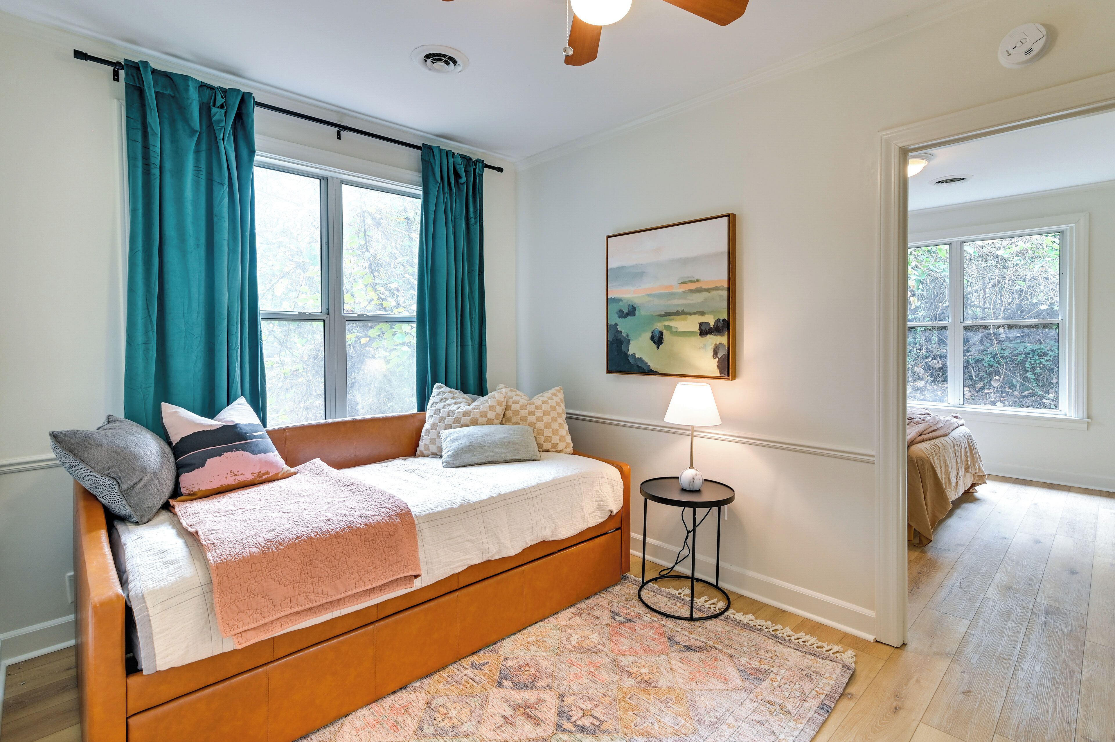 Une chambre avec un lit en bois, des rideaux turquoise, un tableau, une lampe et une vue sur une autre pièce où se trouve un lit.