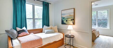Une chambre avec un lit en bois, des rideaux turquoise, un tableau, une lampe et une vue sur une autre pièce où se trouve un lit.