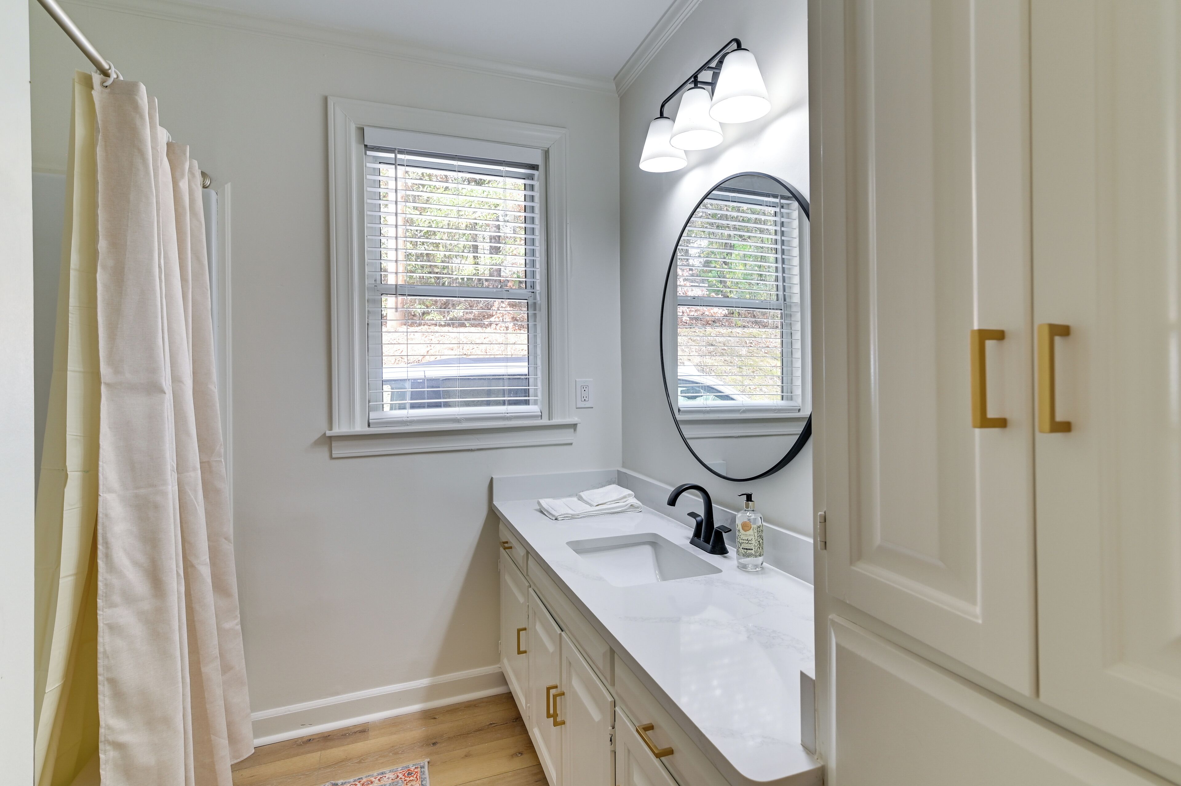 Une salle de bain avec un meuble-lavabo blanc, un grand miroir et une fenêtre avec des stores.
