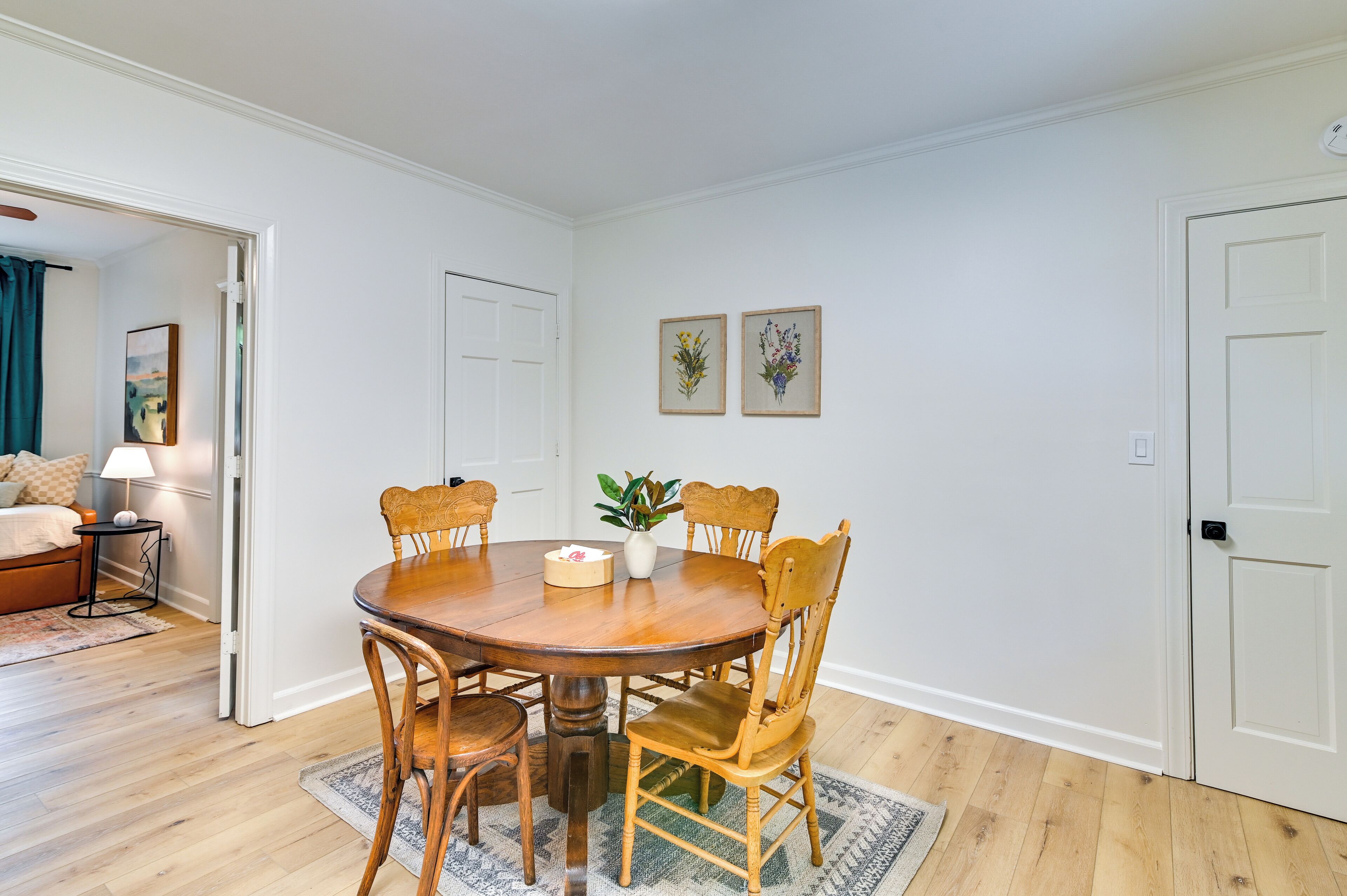 Un coin repas avec une table ronde en bois, six chaises en bois, un vase avec une plante et deux tableaux floraux encadrés accrochés au mur.