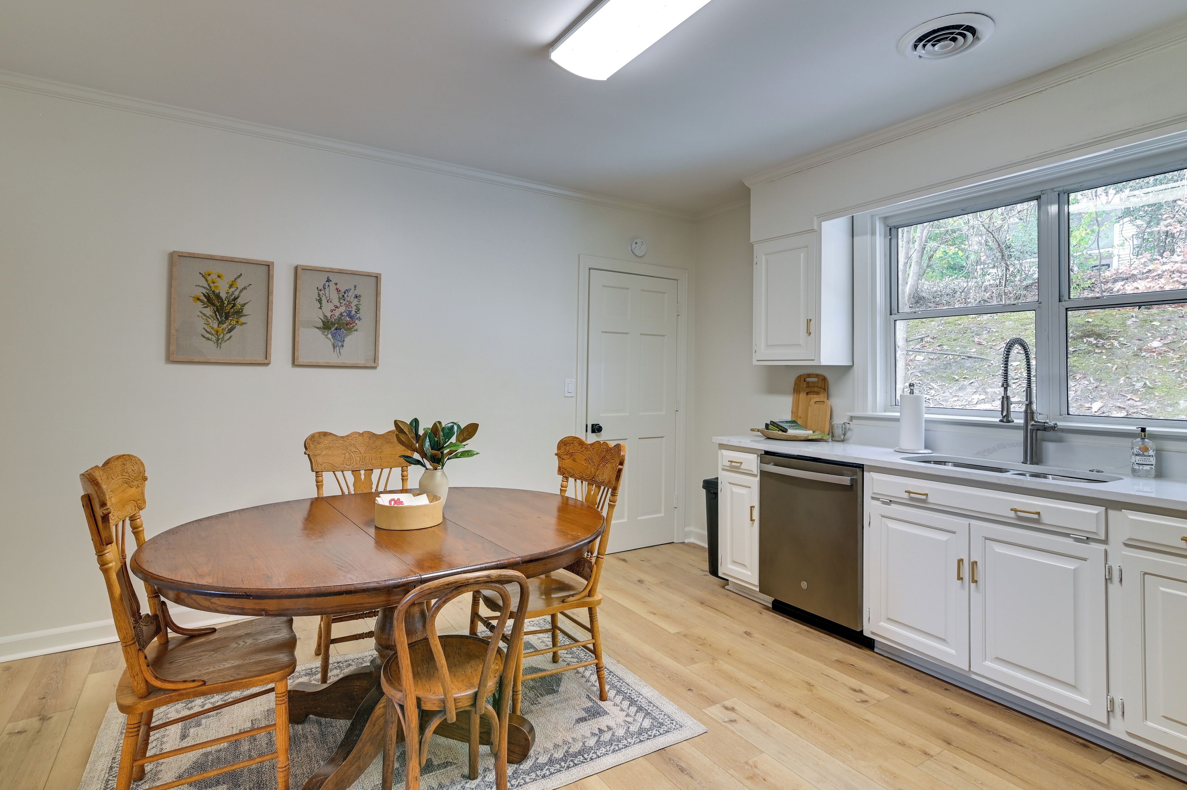 A dining area with a wooden table and chairs, a vase with flowers, and a window with a view of trees.