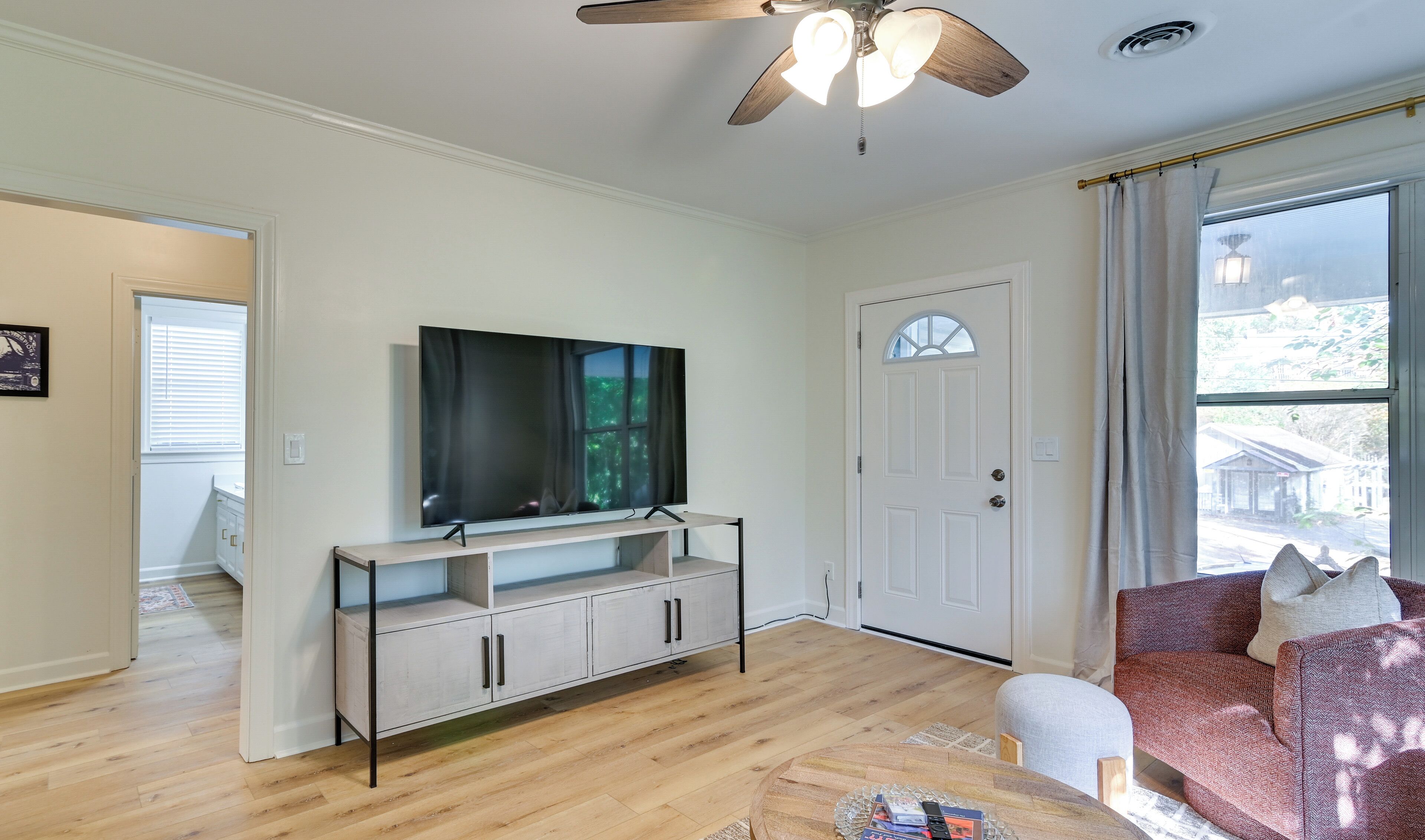 A living room with a TV on a stand, a ceiling fan, a door, and a window with a view of the outside.