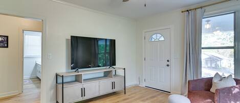 A living room with a TV on a stand, a ceiling fan, a door, and a window with a view of the outside.