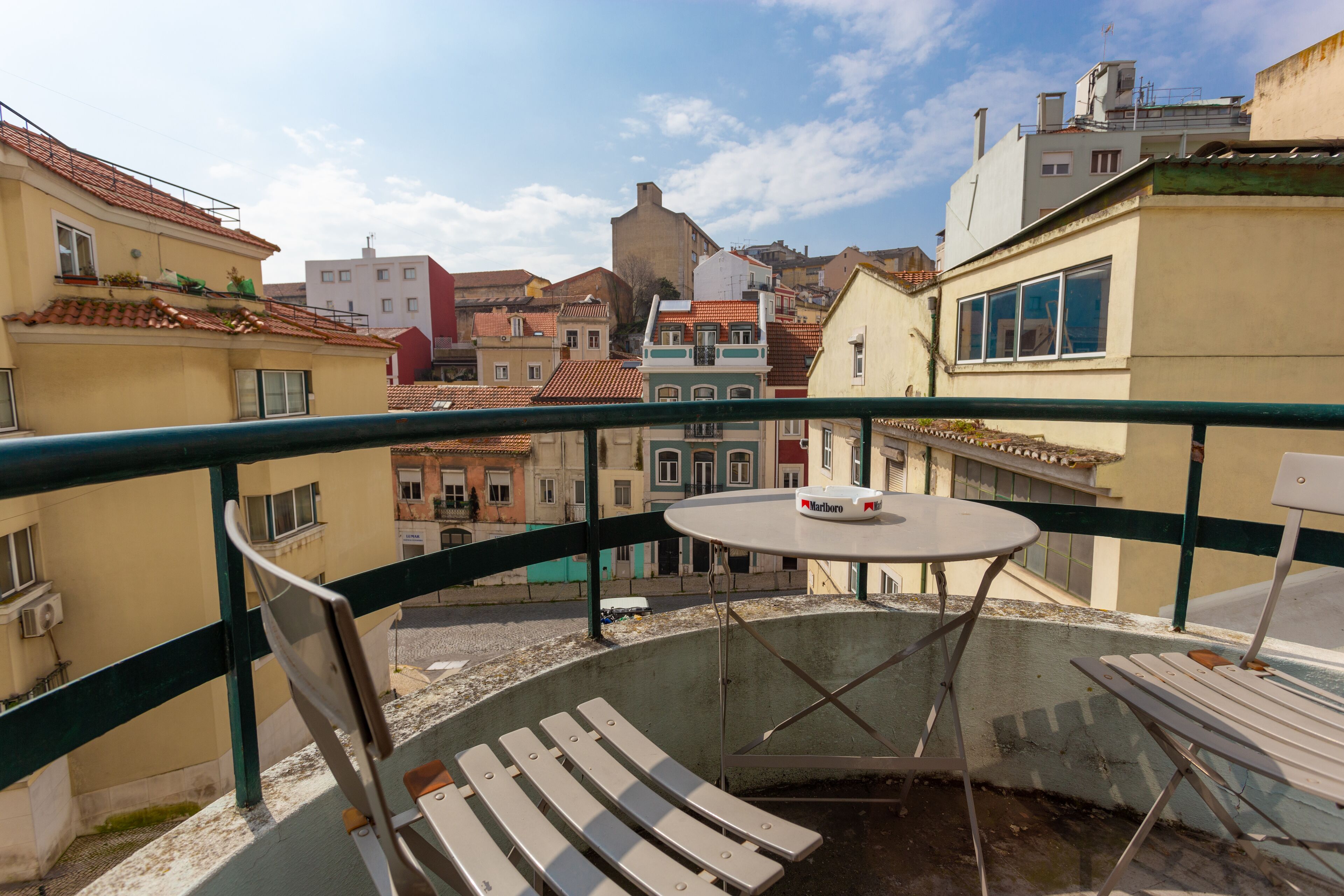 A balcony with a table and chairs, overlooking a cityscape with various buildings.
