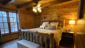Tunnelmallinen hirsihuone, jossa on suuri sänky, puinen pääty, penkki ja kattotuuletin.