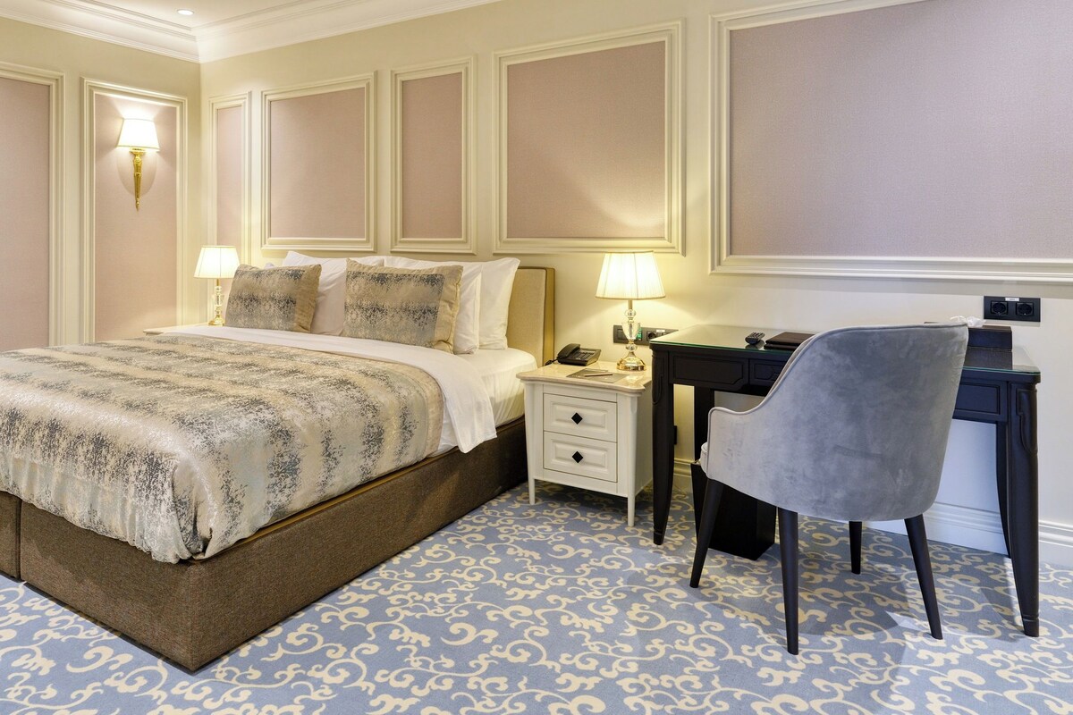 A hotel room with a large bed, a desk with a chair, and a nightstand with a lamp.