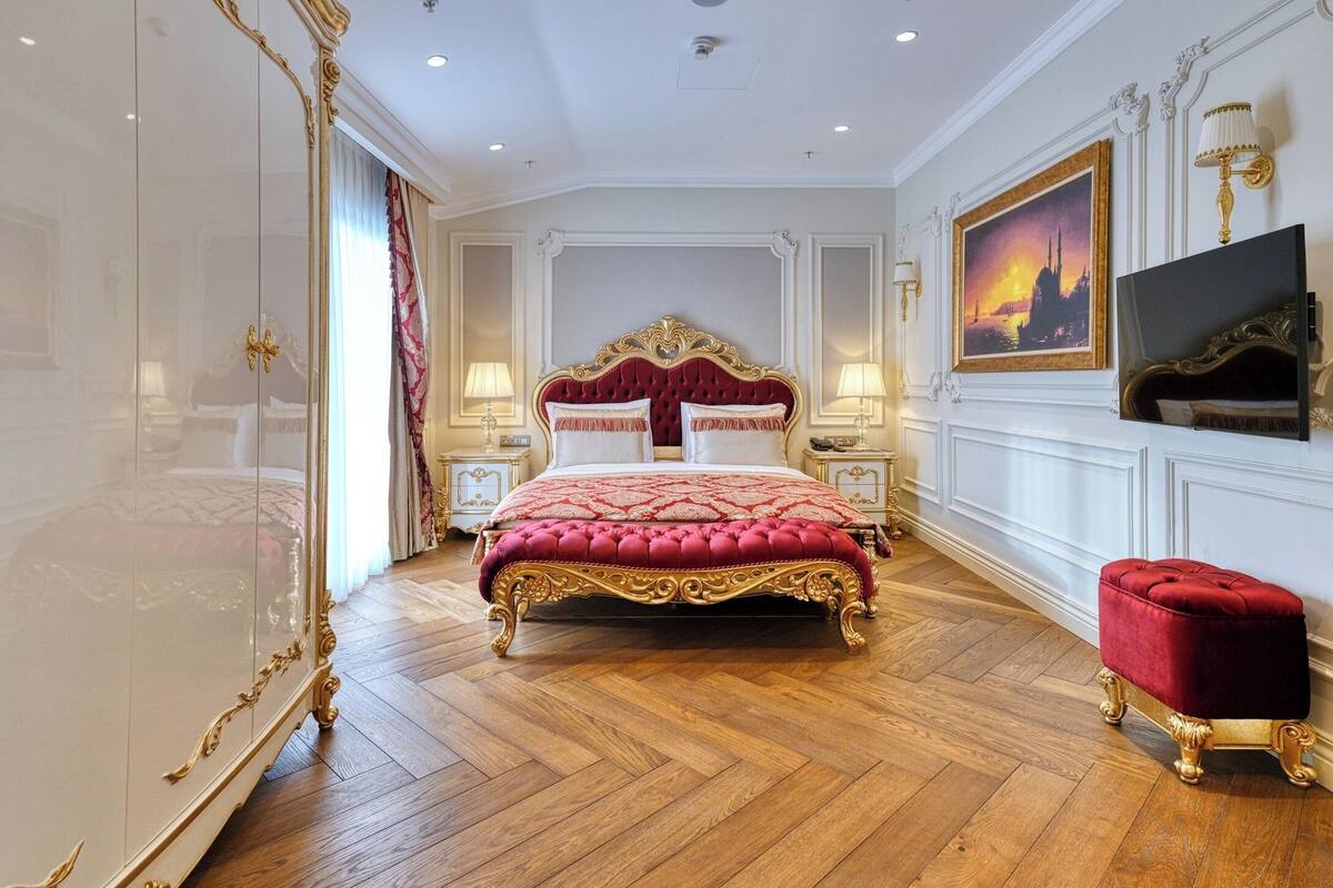 A luxurious bedroom with a large bed, ornate gold detailing, a television, and a red ottoman.