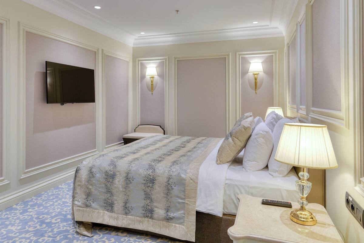 A hotel room with a bed, bedside table, lamp, TV, and wall-mounted light fixture.