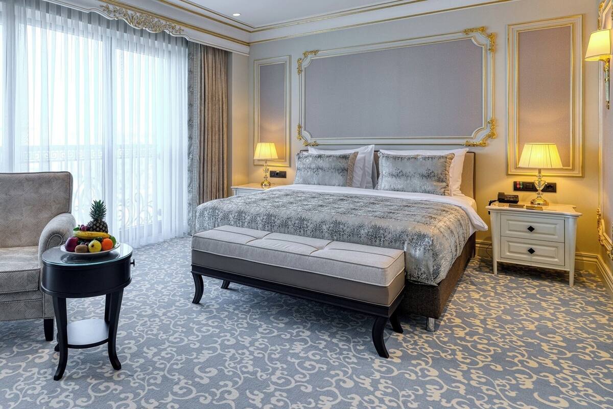 A hotel room with a large bed, a chair, a small table with fruit, and a nightstand with a lamp.