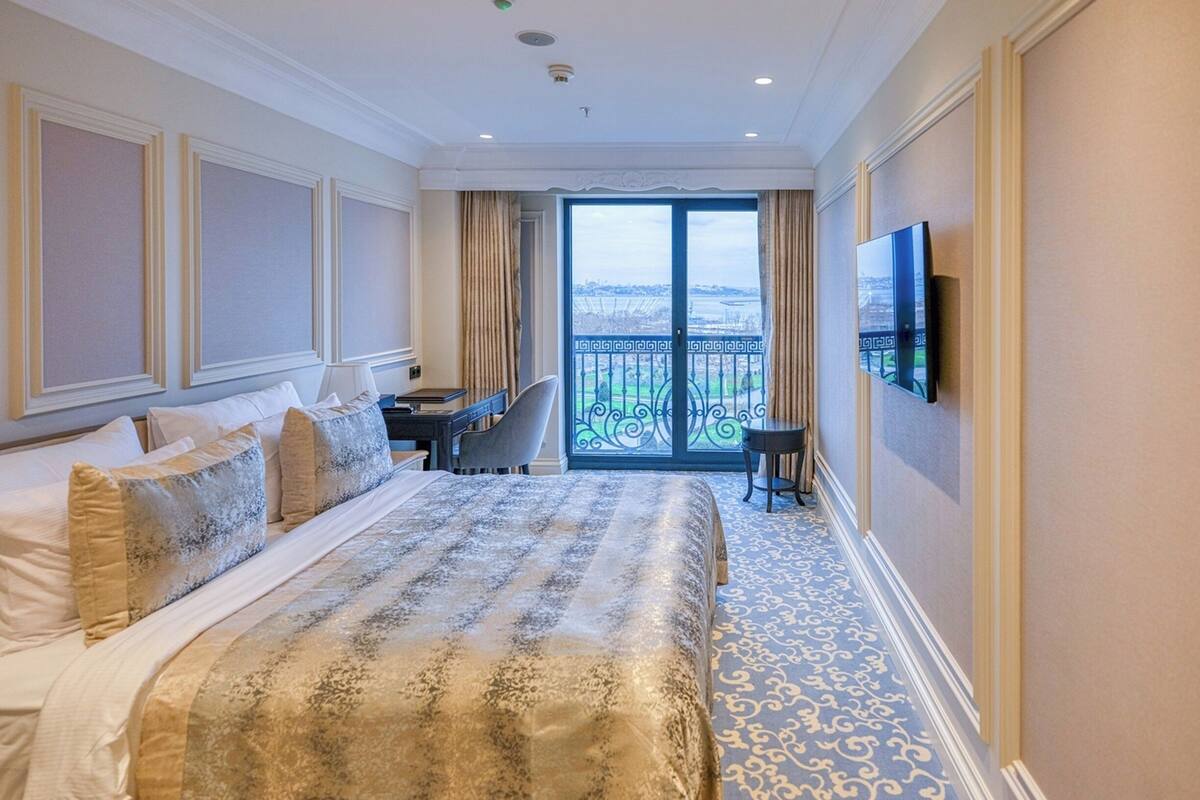 A hotel room with a large bed, a desk, a chair, a TV, and a balcony with a view.
