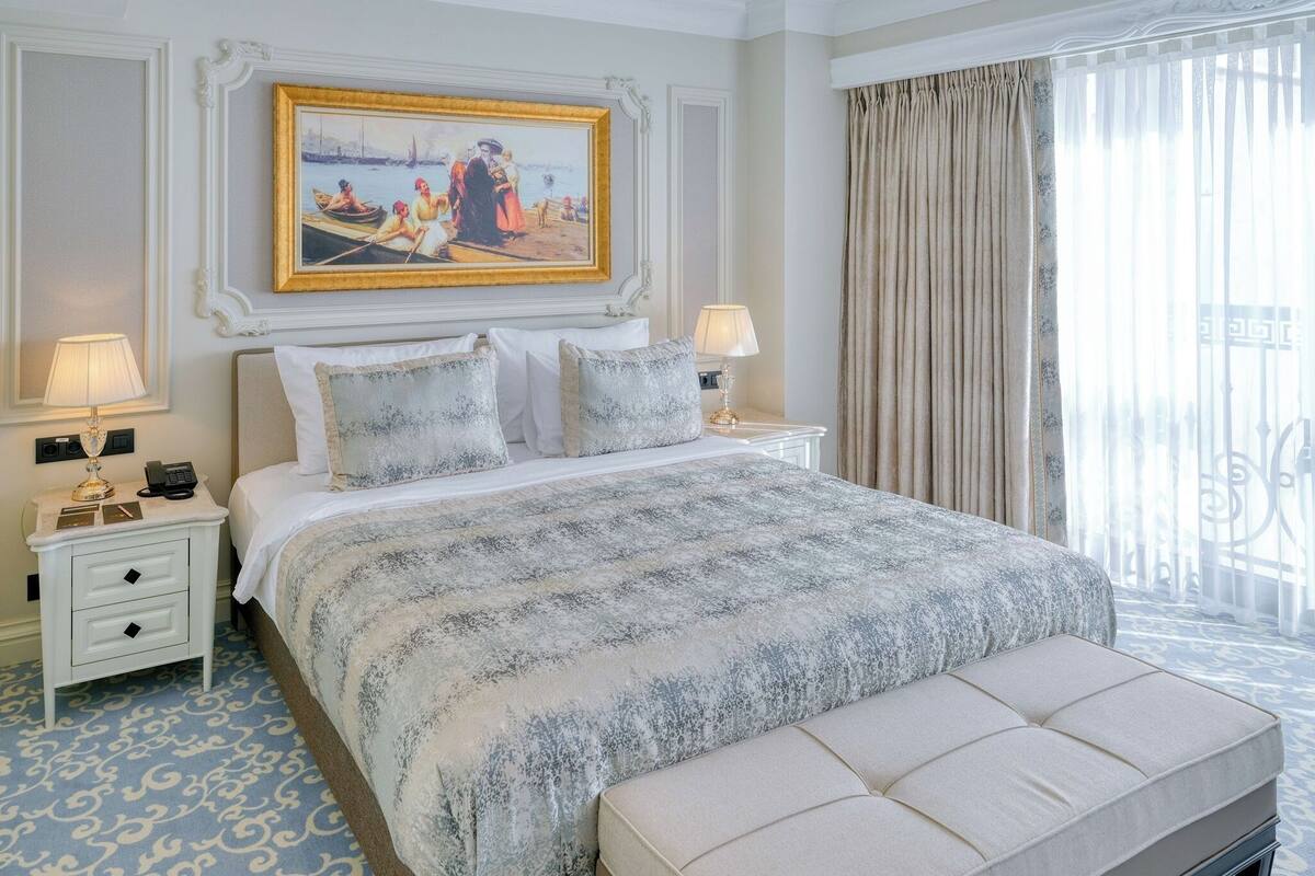 A hotel room with a large bed, two bedside tables with lamps, a painting on the wall, and a view of the outdoors through a window with curtains.