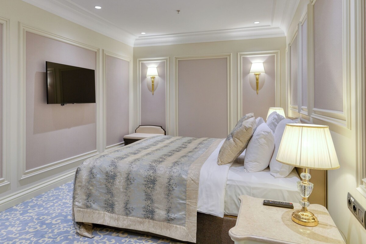 A hotel room with a bed, bedside table, lamp, TV, and wall-mounted light fixture.