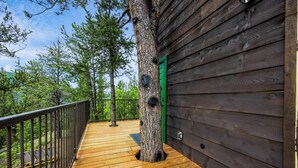 Une terrasse en bois avec une rambarde en métal, une porte verte et le tronc d’un arbre au centre.