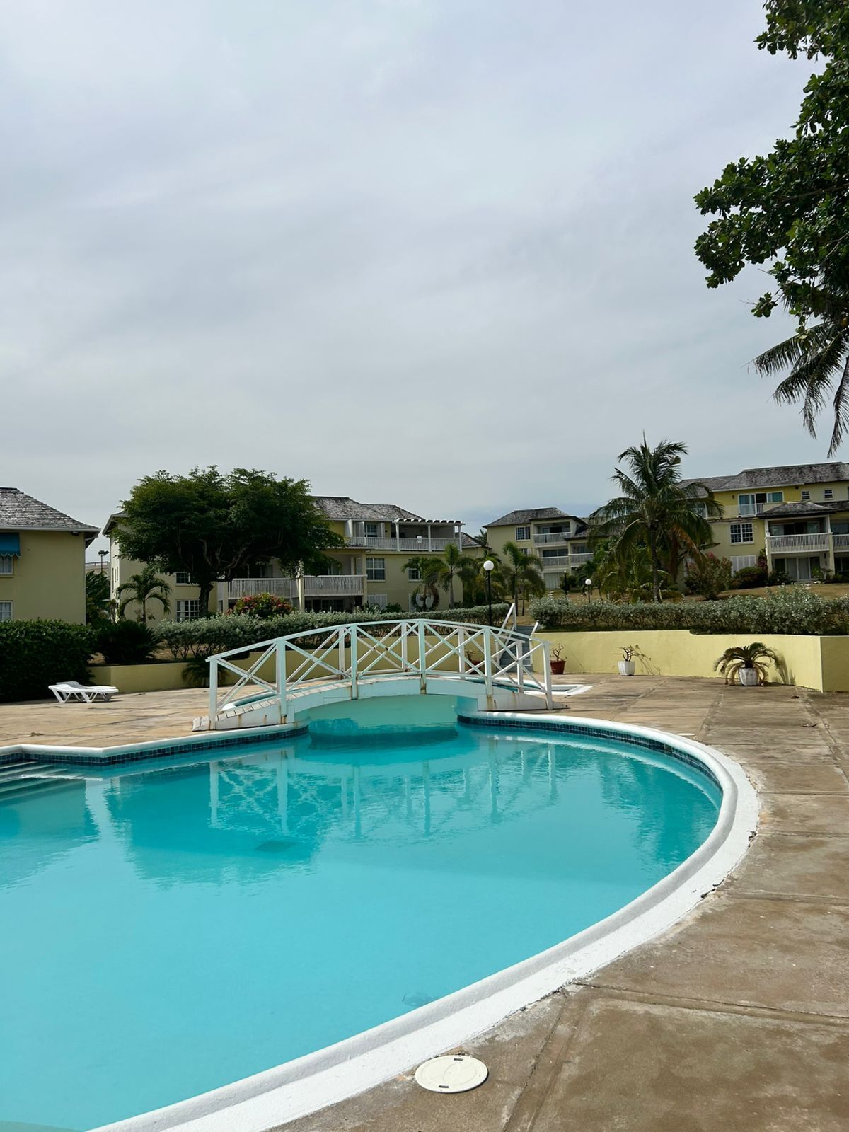 A swimming pool with a white bridge, surrounded by buildings with balconies and palm trees.