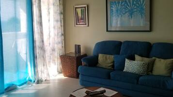 A living room with a blue sofa, a woven ottoman, a ceiling fan, and an air conditioning unit.