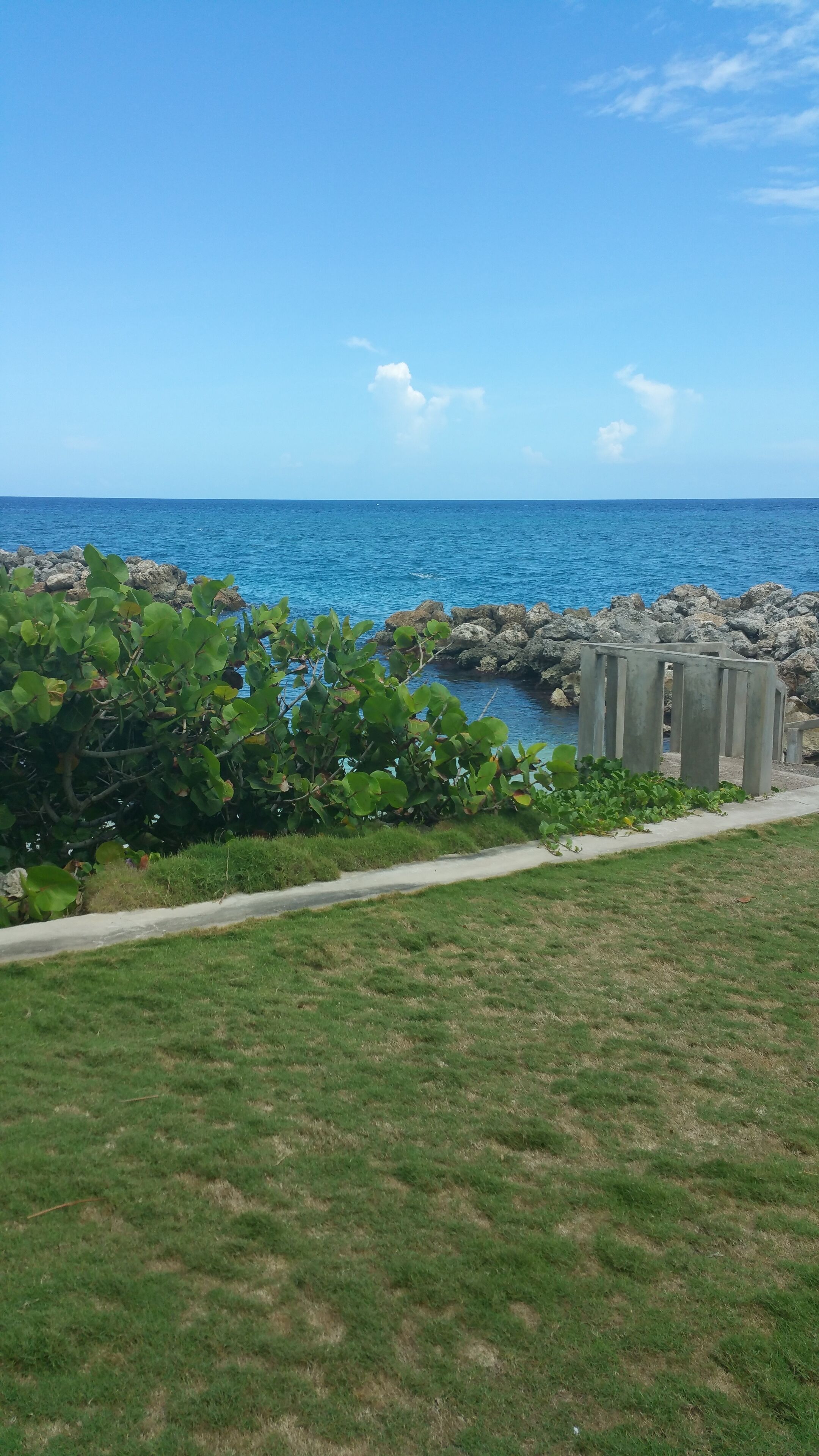 A coastal view with a grassy area, a concrete pathway, and rocky outcrops.