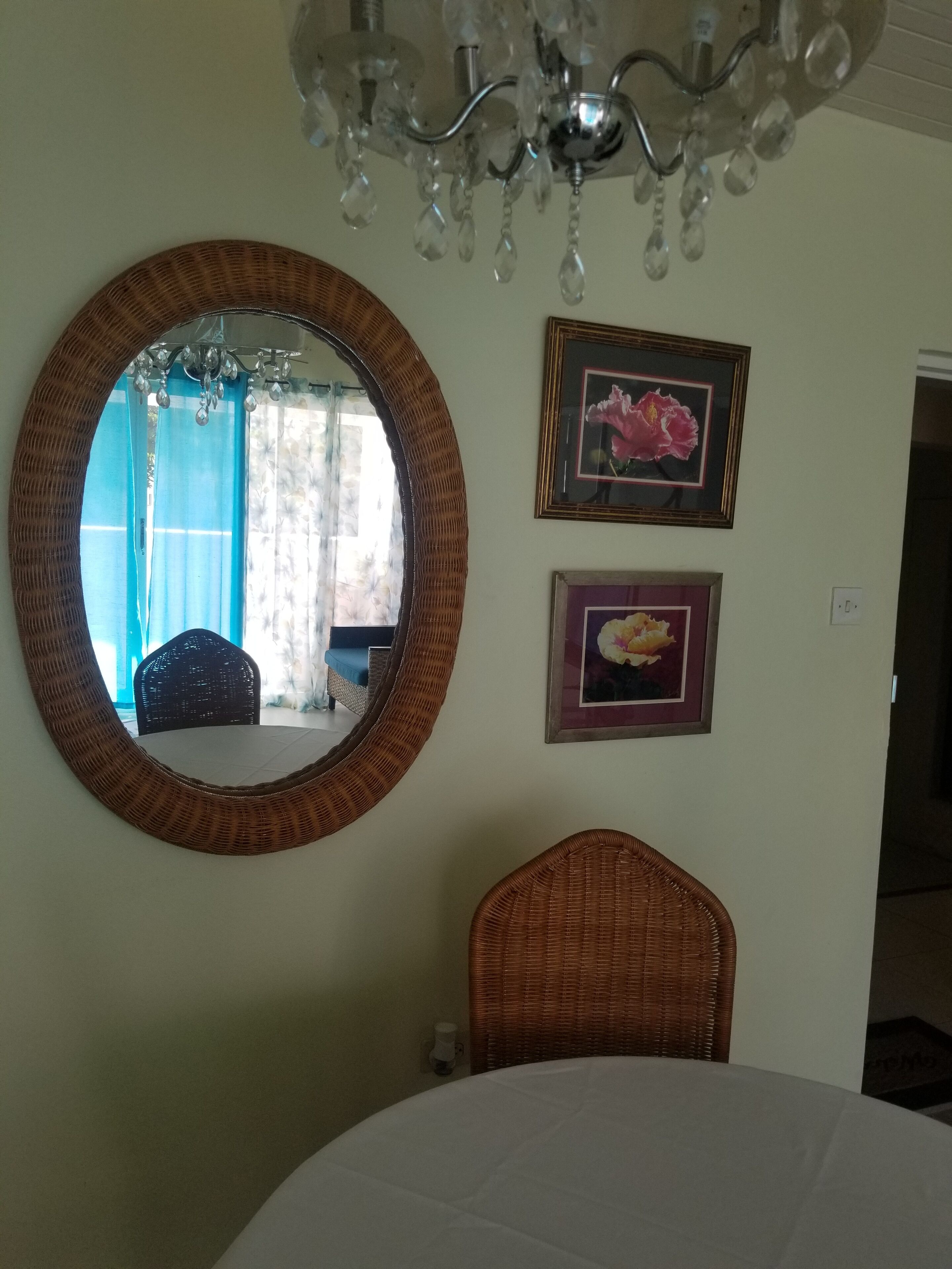 Een kamer met een ronde spiegel, twee ingelijste foto's, een kroonluchter en een rieten stoel.