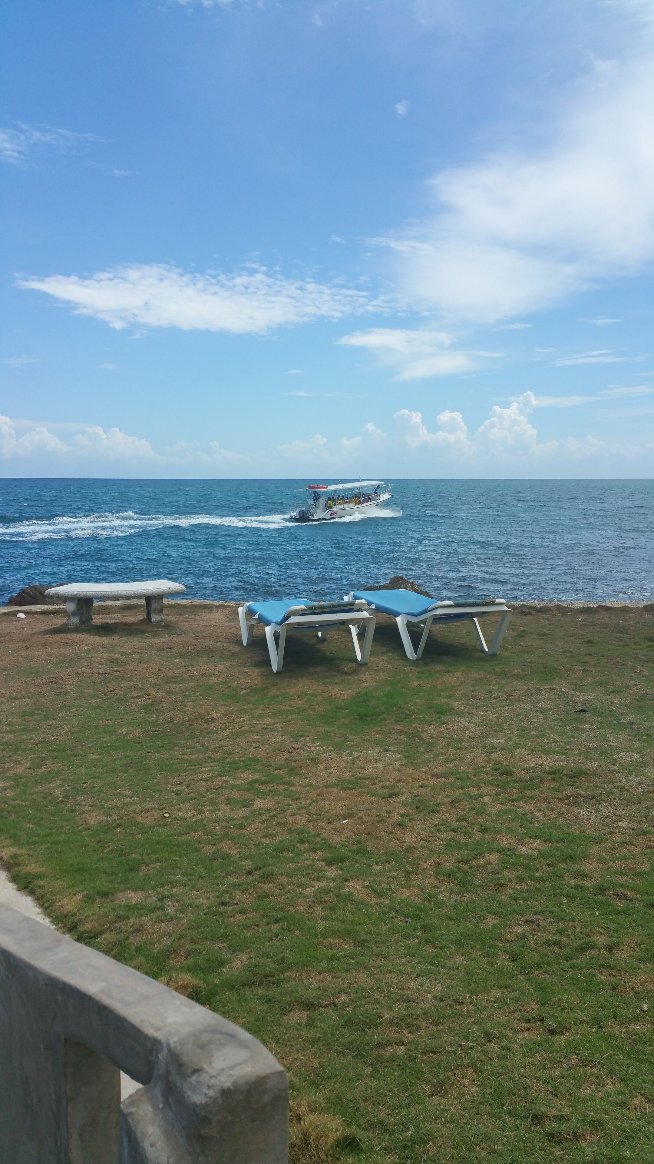 Un espacio cubierto de césped con dos sillas de descanso y un banco de piedra, con vista al mar tranquilo y un barco a lo lejos.