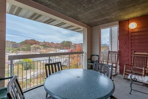 Un balcón con mesa y sillas, vista de un paisaje con árboles, y una pared roja con una lámpara.