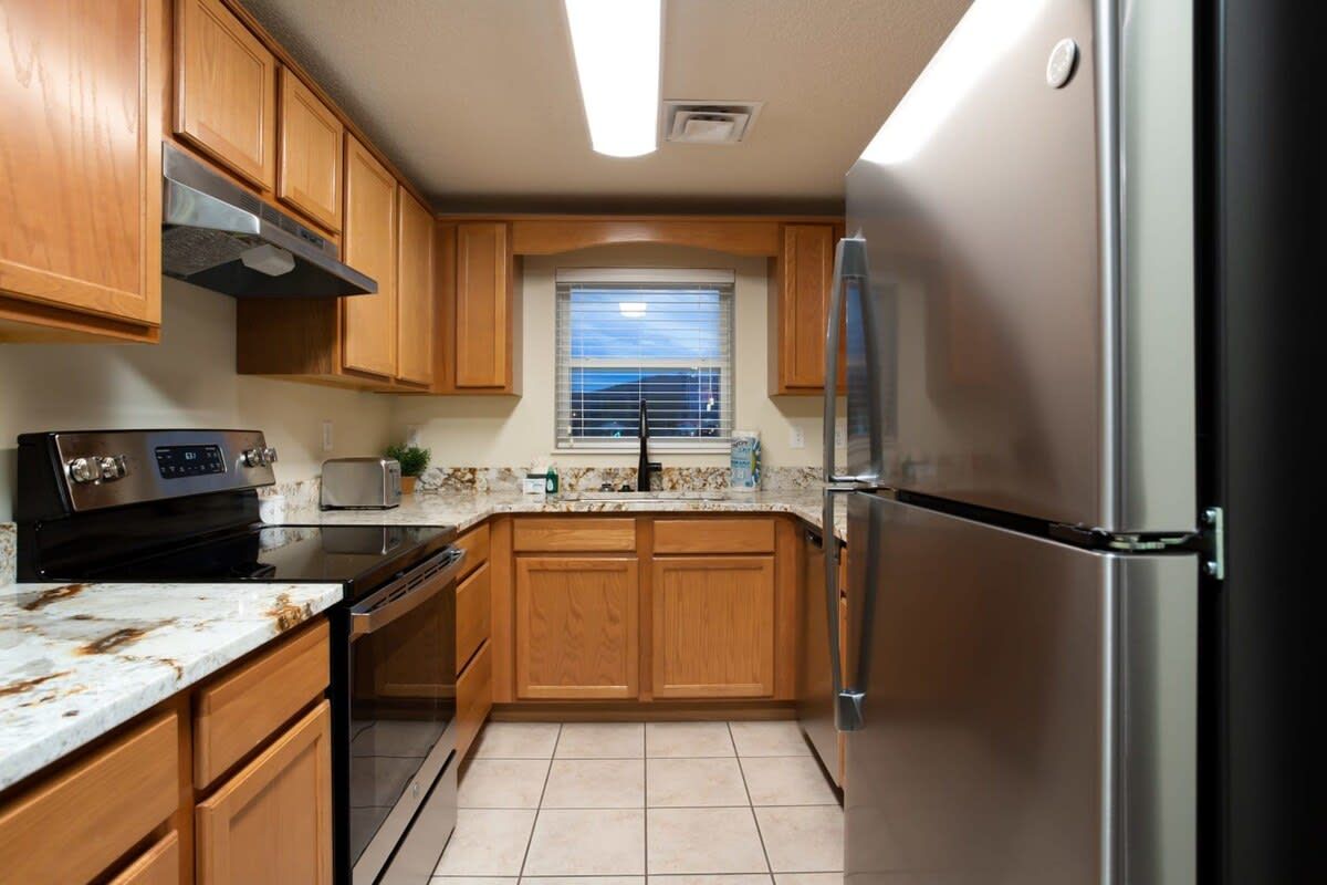 A compact kitchen with wooden cabinets, a stainless steel refrigerator, a built-in oven, and a window with blinds.