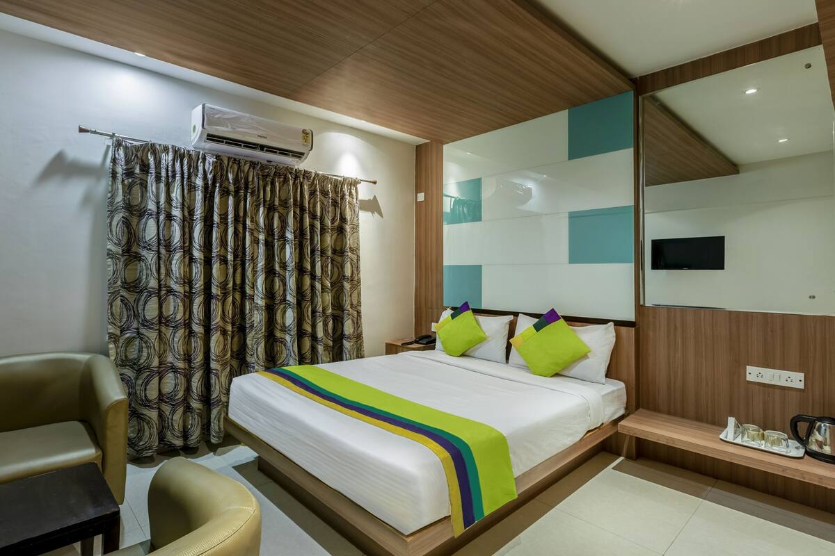 A hotel room with a bed, a desk, a chair, an air conditioning unit, a television, and a mirror.