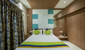 Una habitación de hotel moderna con una cama grande, una mesita de noche con un teléfono, y una pequeña mesa con una tetera y vasos.