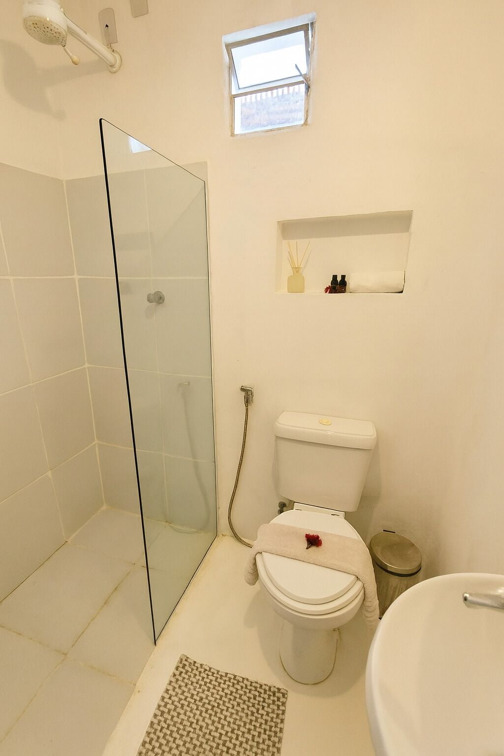 Un baño con cabina de ducha de vidrio, un inodoro con un pequeño estante encima y una ventana.