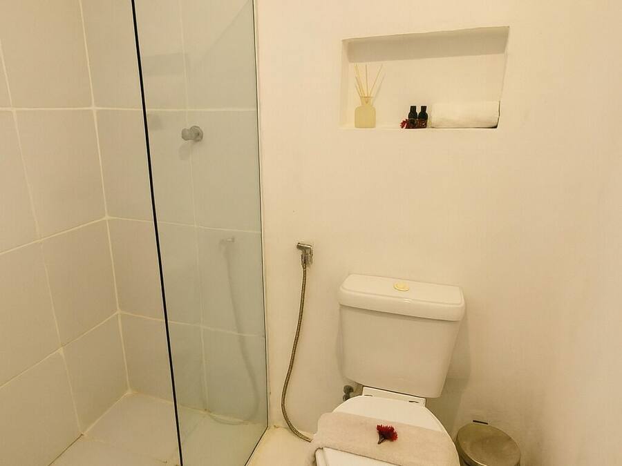 Um banheiro com box de vidro, vaso sanitário com prateleira pequena acima e uma janela.