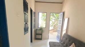 A spacious room with a grey sofa, a small table with a plant, and a view of greenery outside. - Pousada Vilarejo (Porto Seguro)