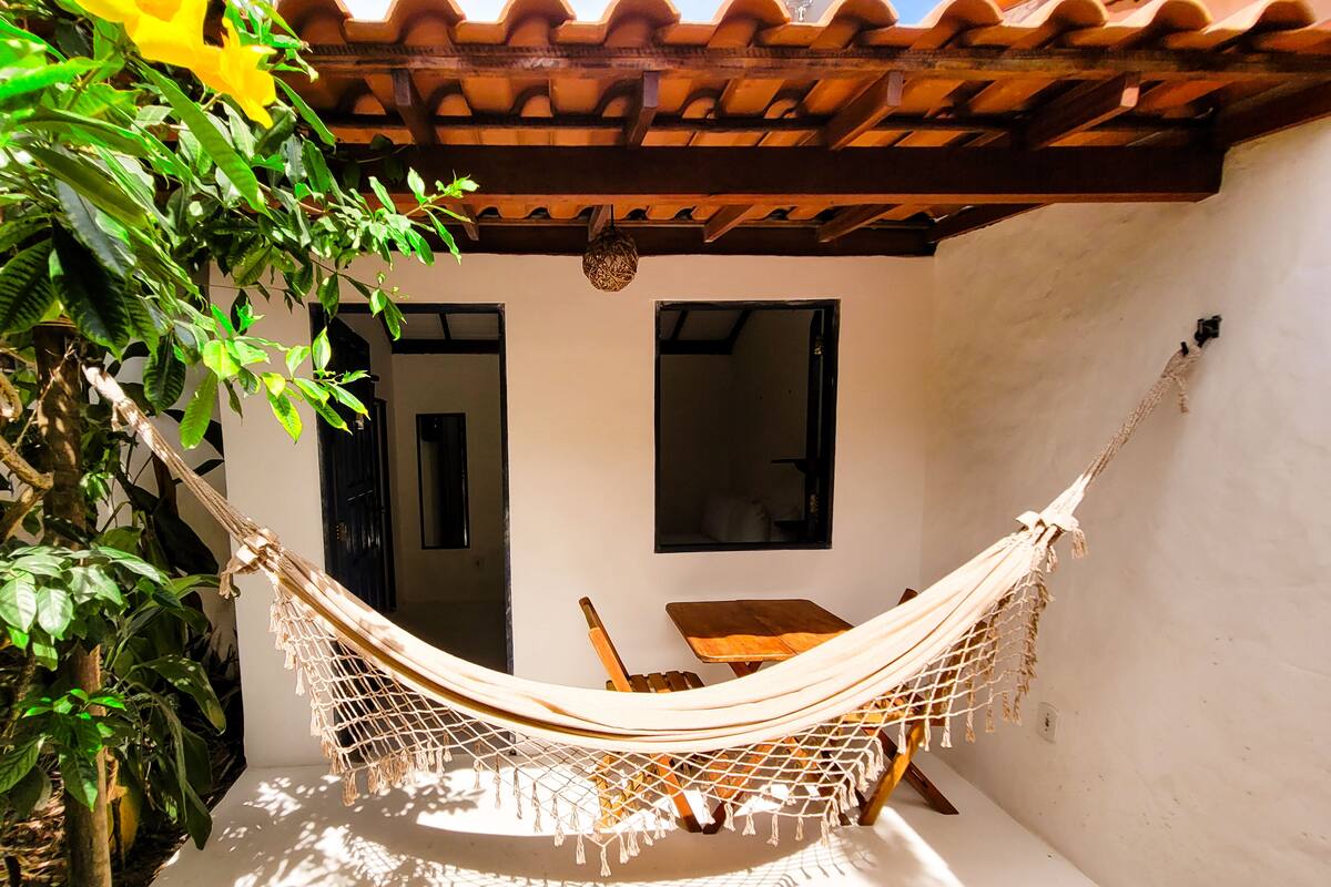 A hammock set up between two wooden chairs under a tiled roof, with a white wall and a window in the background.