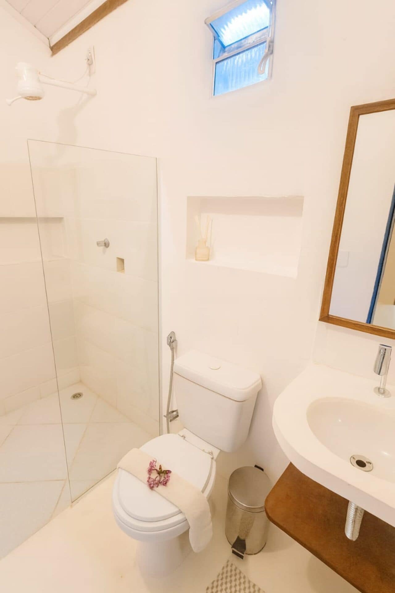 Un baño moderno con cabina de ducha de vidrio, un inodoro decorado con una flor, un lavamanos con espejo y un pequeño estante con una vela.