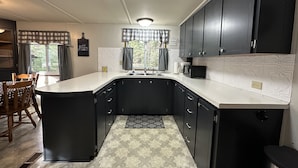 A compact kitchen with dark cabinets, a white countertop, a built-in sink, and a window with curtains.