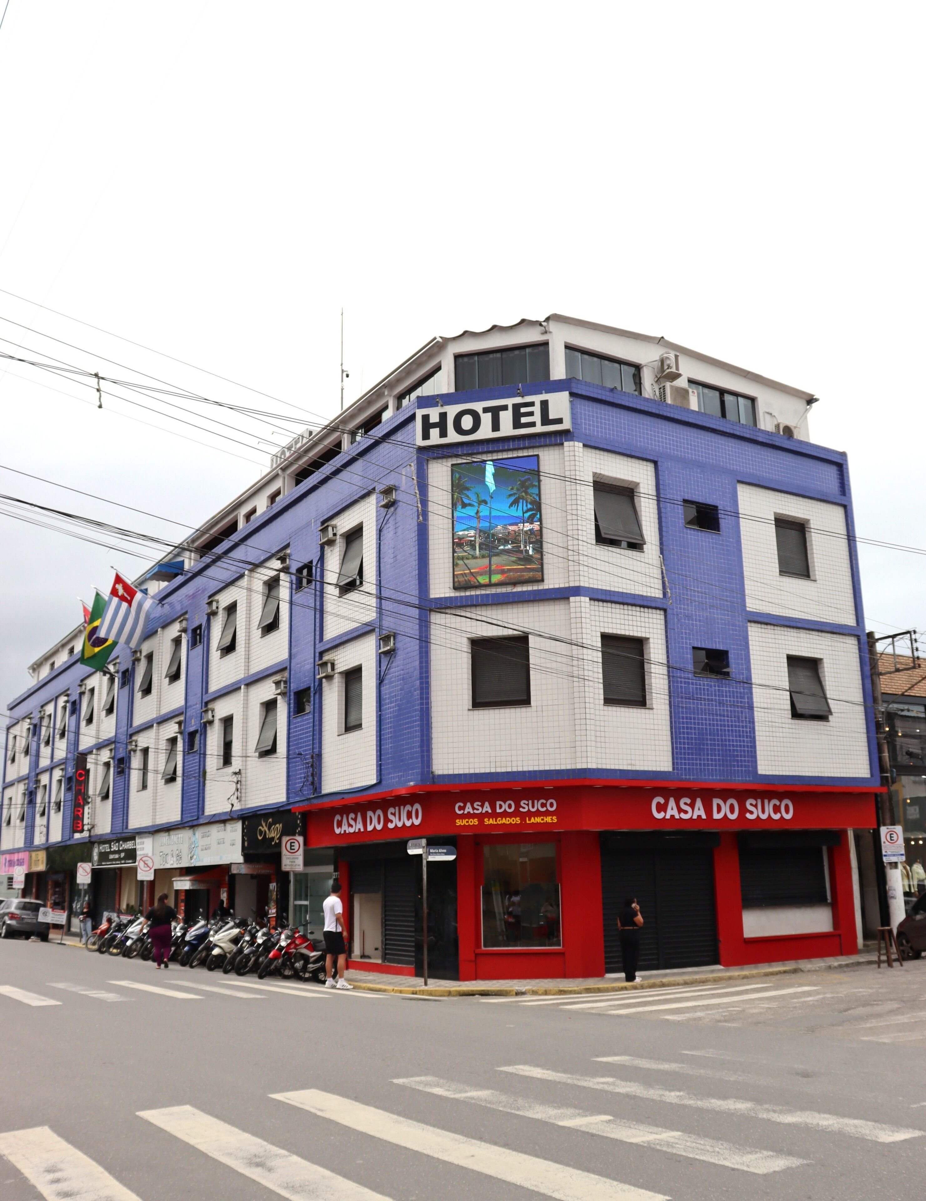 Un bâtiment d’hôtel avec une enseigne indiquant qu’il s’agit d’un hôtel, une devanture de magasin avec une enseigne pour « Casa do Suco », et plusieurs motos garées.