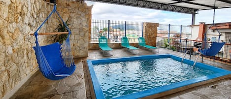 An outdoor swimming pool with stone walls and a metal roof, surrounded by lounge chairs and a hanging chair.