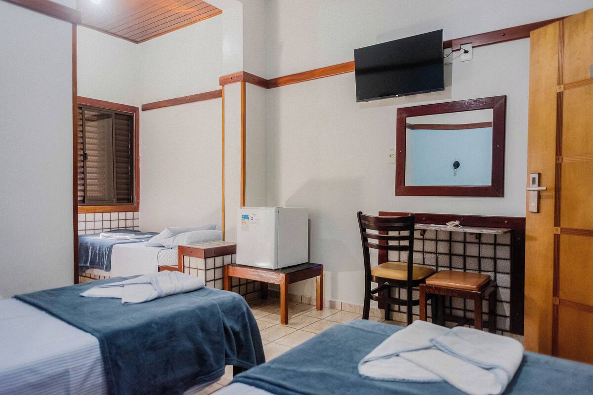 A hotel room with two beds, a television, a mirror, a refrigerator, and a window with blinds.