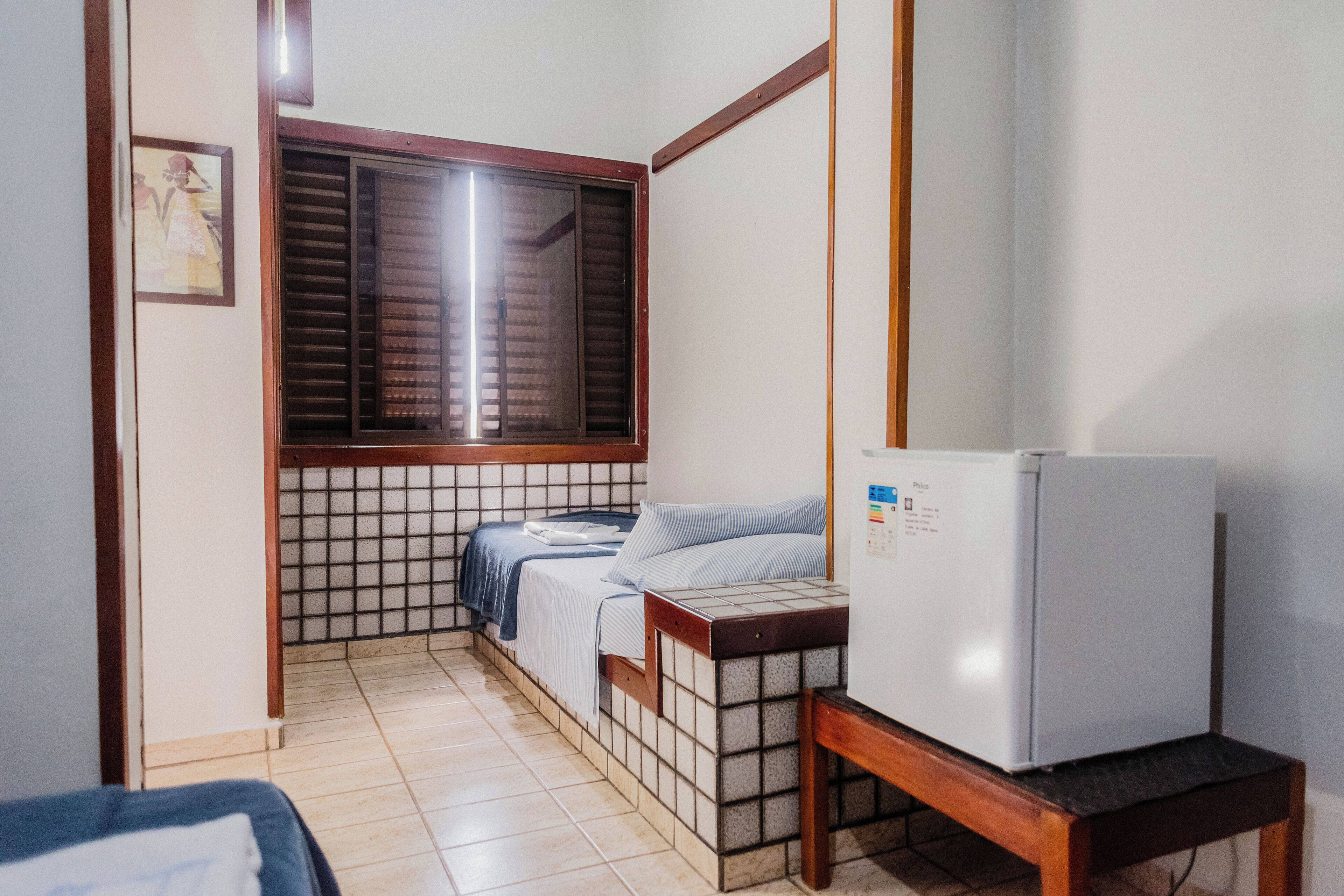 A small room with a bed, a window with blinds, a refrigerator, and a tiled floor.