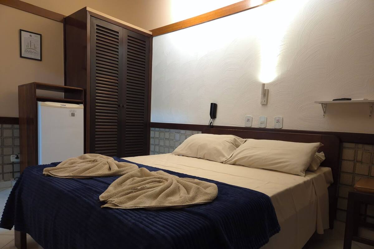 A hotel room with a bed, a wardrobe, a small refrigerator, a bedside table, and a wall-mounted phone.