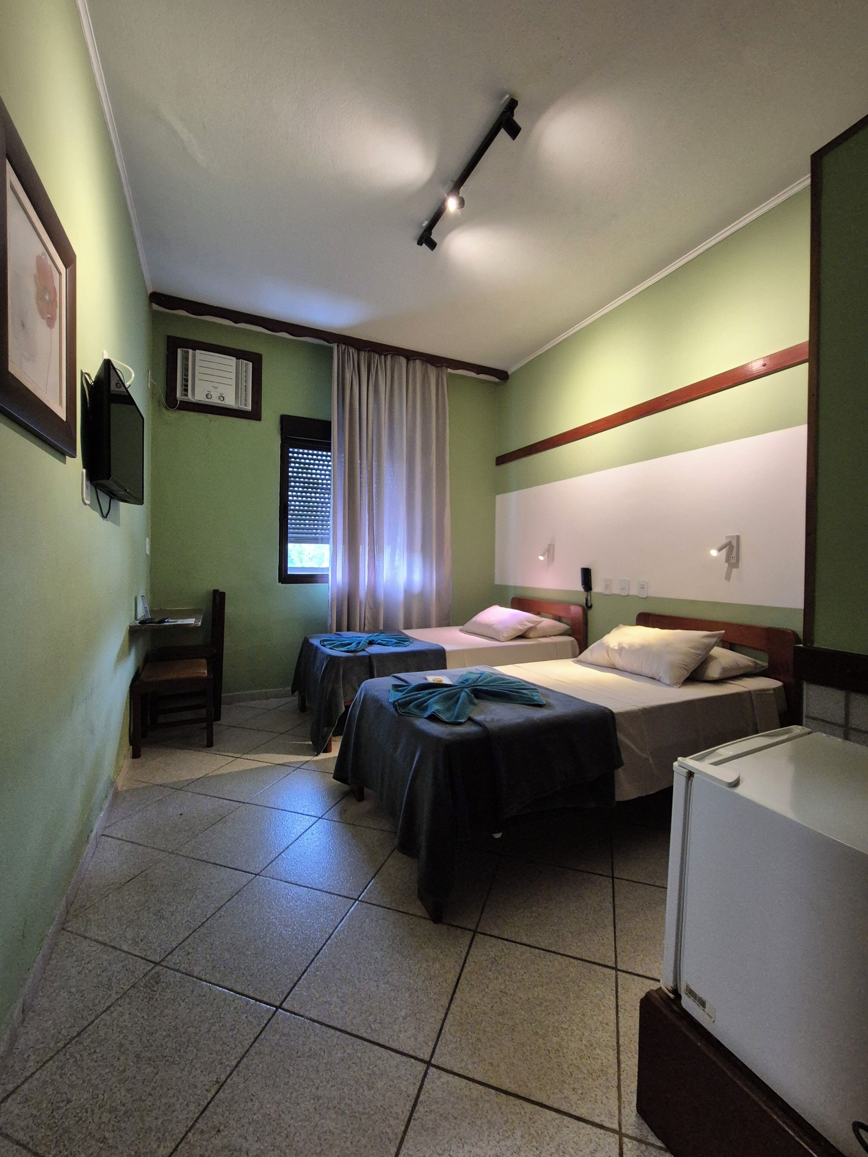 Ein Hotelzimmer mit zwei Betten, einem Schreibtisch, einem Stuhl, einem Fernseher und einem Vorhangfenster.