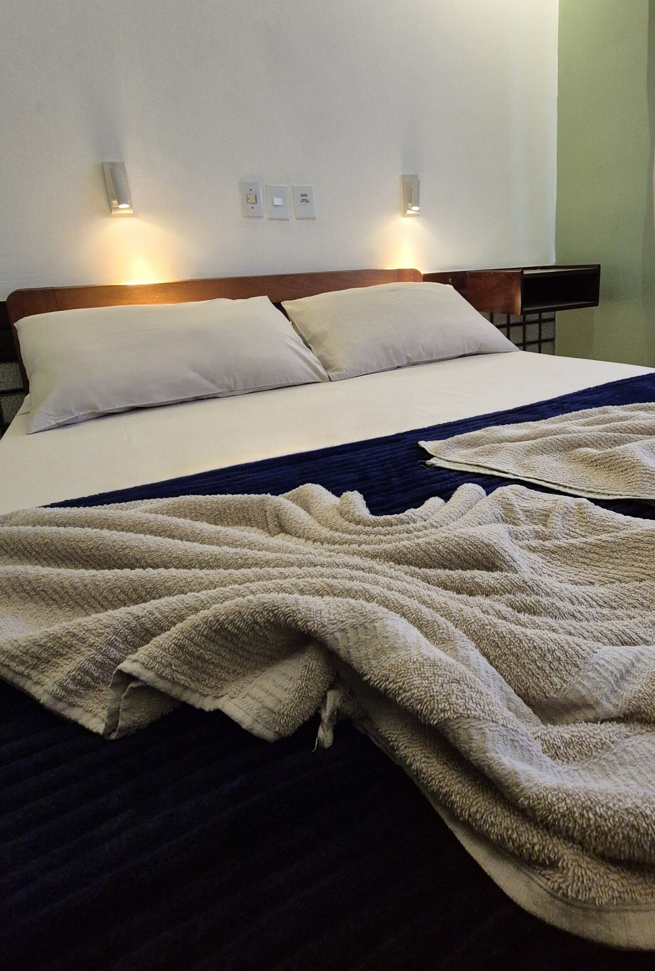 Un letto con copripiumino bianco e blu, una coperta beige e due luci fissate al muro.