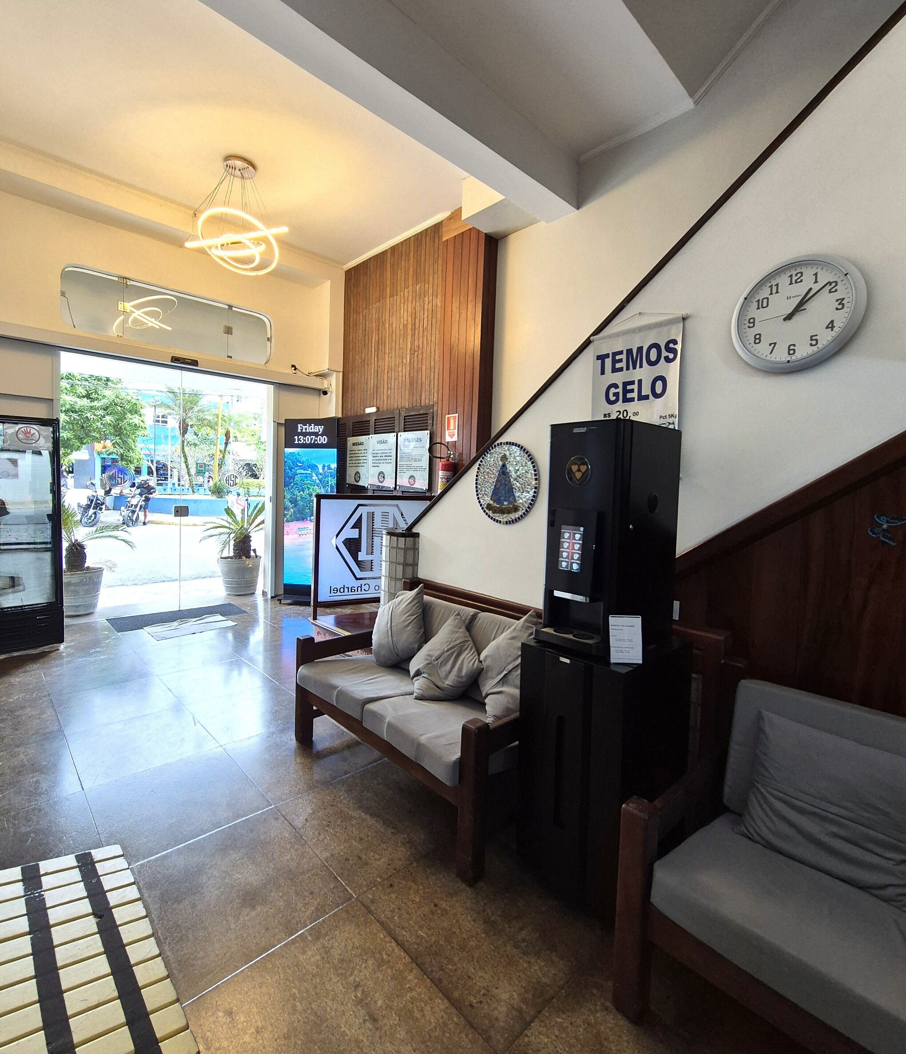 A reception area with a sofa, a clock, a sign reading 'TEMOS GELO,' and a digital screen displaying information.