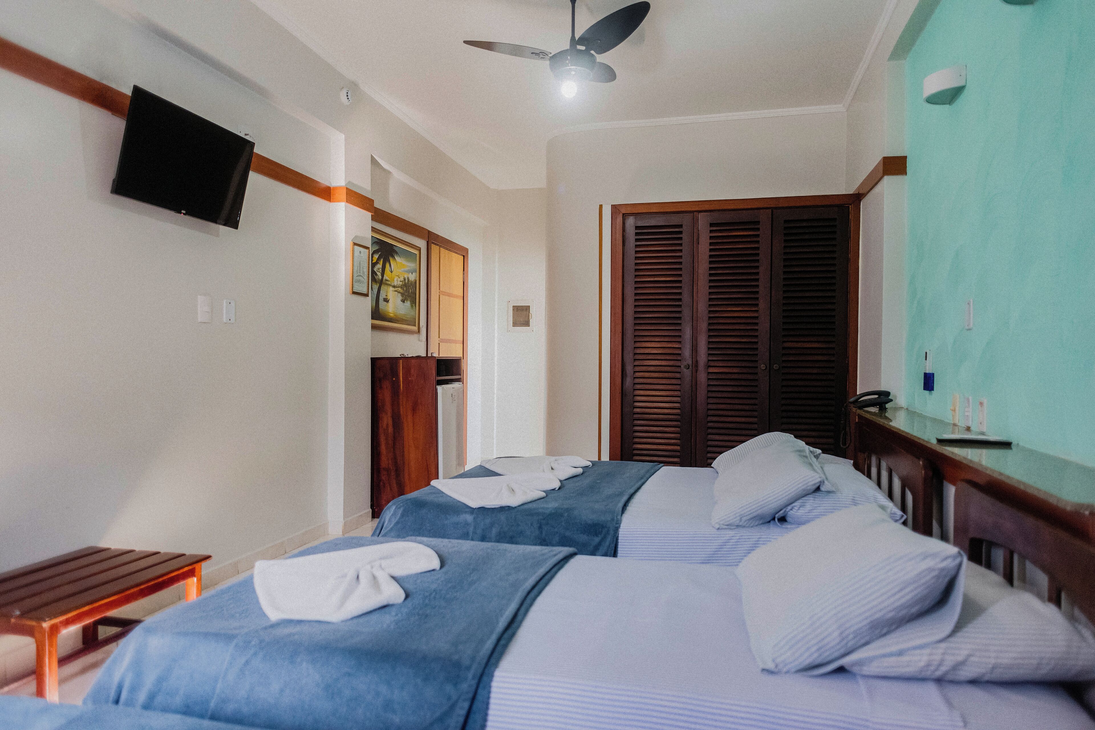 A hotel room with two beds, a television mounted on the wall, a ceiling fan, and a wooden door.