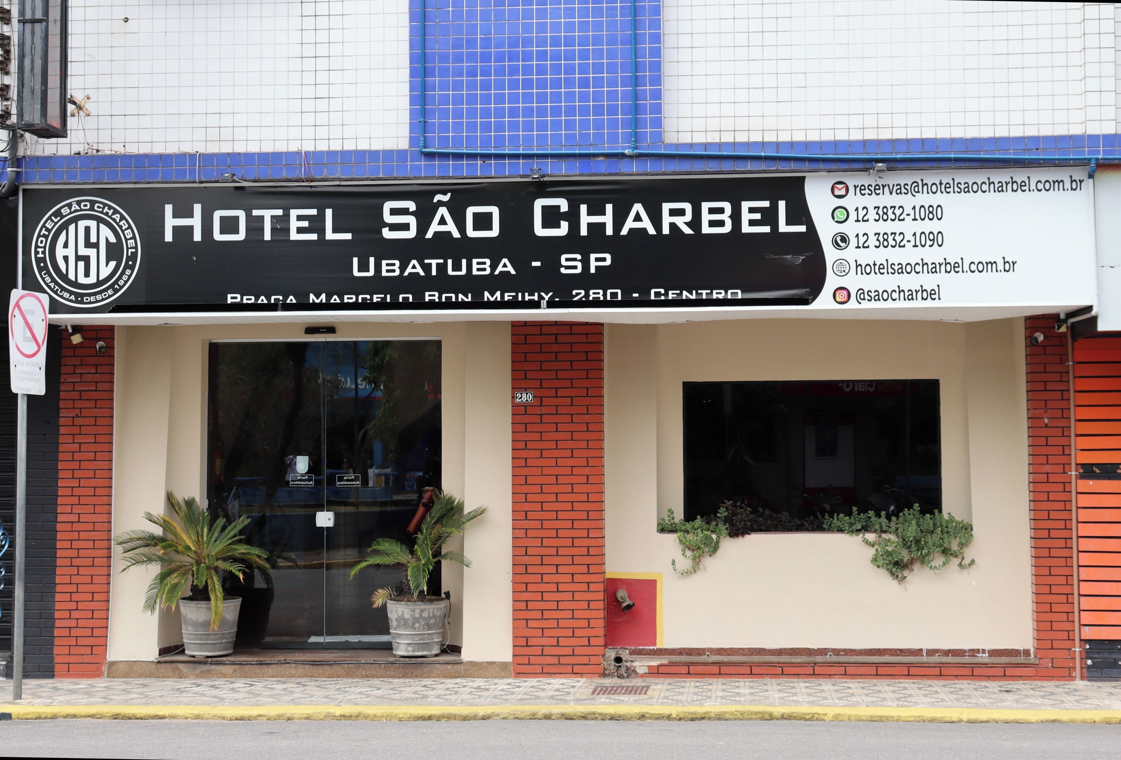 Un hotel con un cartel que muestra el nombre 'HOTEL SÃO CHARBEL' e información de contacto.