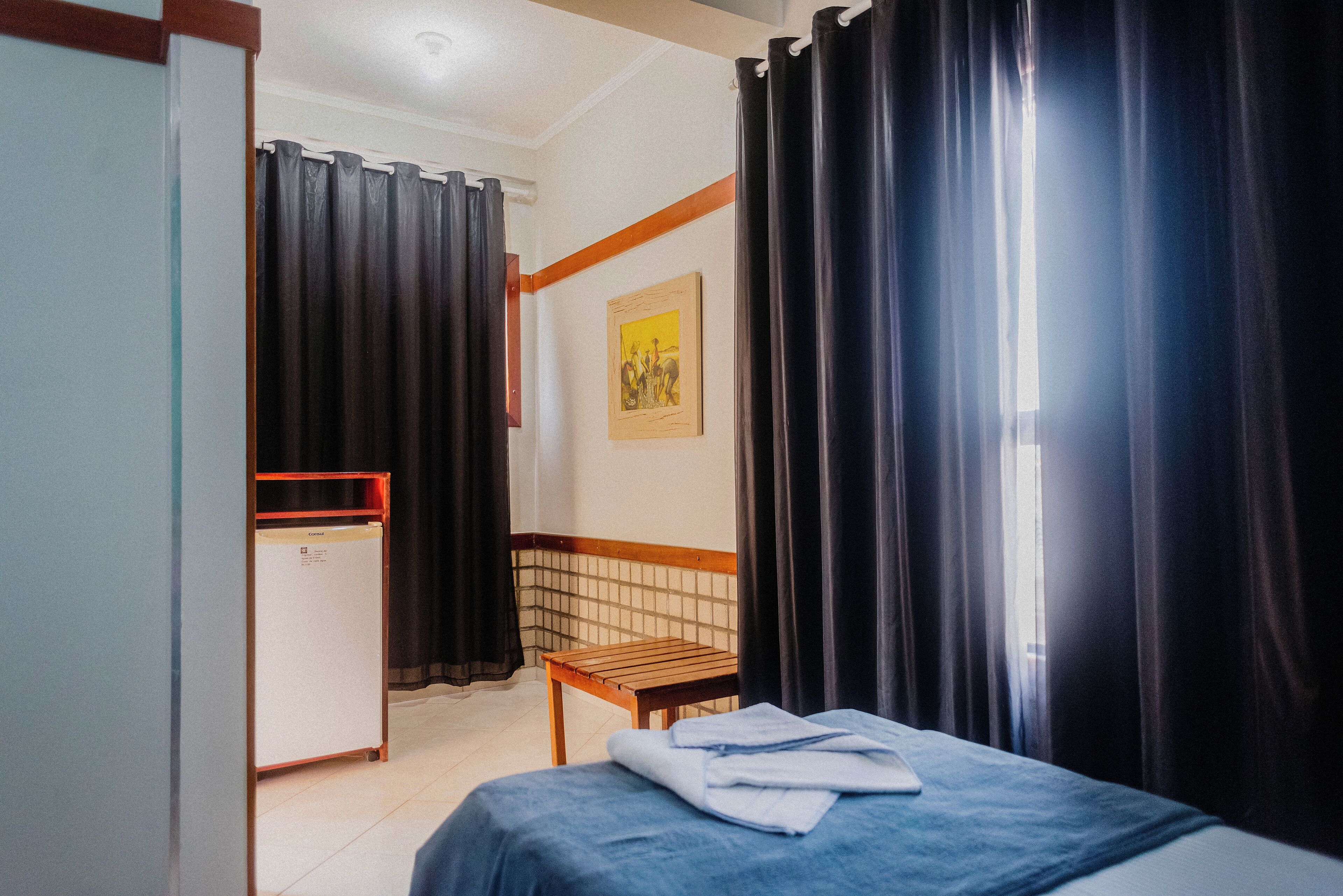 Quarto de hotel com cama, uma cadeira de madeira, escrivaninha com computador e janela com cortinas.