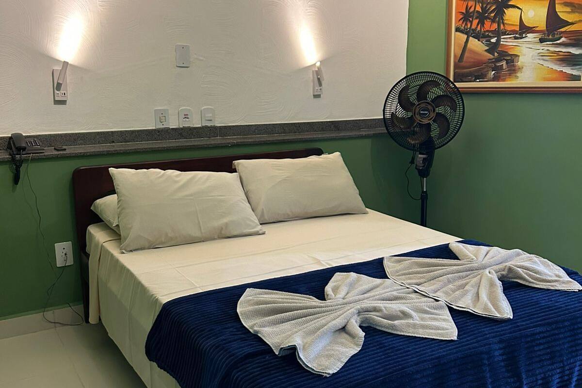 A hotel room with a bed, a fan, and a painting on the wall.