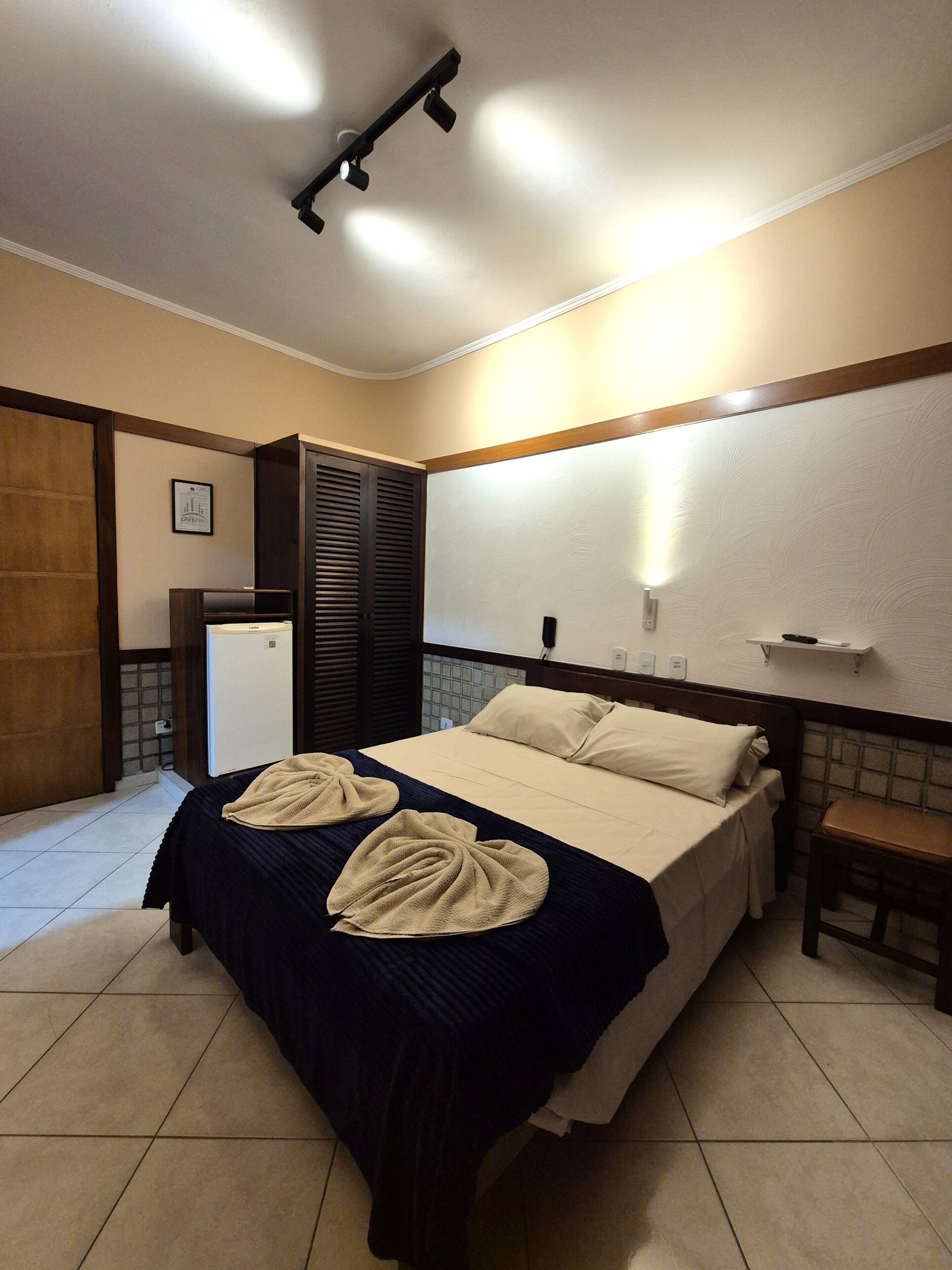 A hotel room with a bed, a desk, a chair, a wardrobe, and a small refrigerator.