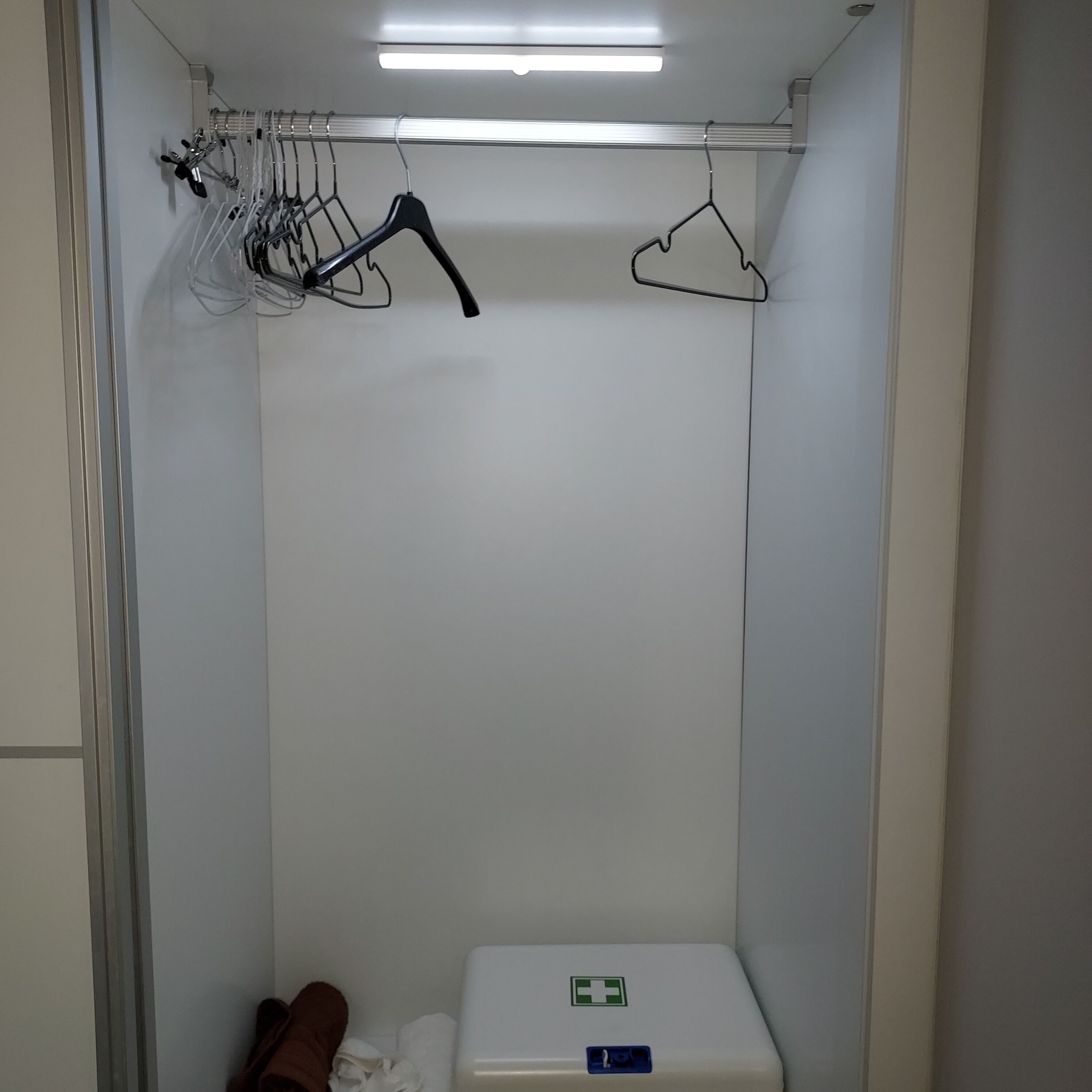 A small, enclosed space with a white wall, a metal rack with hangers, and a white container with a green cross symbol.