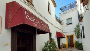 Eine enge Kopfsteinpflasterstraße mit Häusern an beiden Seiten. Ein rotes Markisenvorzelt mit dem Schriftzug "Banos Arabes de Cordoba" ist sichtbar. Im Vordergrund befinden sich Blumentöpfe und ein kleiner Baum.